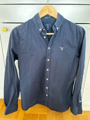 Mörkblå skjorta från GANT storlek 158-164 - Snygg mörkblå skjorta från GANT med klassisk button-down krage och vita knappar. Skjortan har broderad logga på bröstet och är tillverkad i mjuk bomull. Perfekt för dig som gillar stilrena och tidlösa plagg. Storlek 158-164