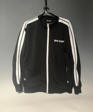 Palm Angels zip - Svart zip-tröja från Palm Angels med vita ränder längs ärmarna och logotyp på bröstet. Tröjan har ståkrage, långa ärmar och två fickor med dragkedja. Snygg sportig look och tillverkad i polyester för en skön känsla.
