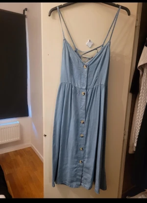 Ljusblå jeansklänning från H&M Divided - Säljer en ljusblå jeansklänning från H&M Divided i storlek 36. Klänningen har smala axelband, snygg snörning i ryggen och knappar framtill hela vägen ner. Materialet är mjukt denim och modellen är lång med en avslappnad passform. 