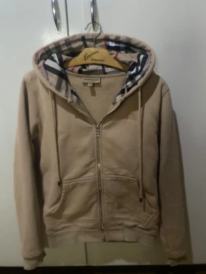 Burberry hoodie    - Säljer en beige hoodie från Burberry i storlek S. Tröjan har dragkedja, snörning och den klassiska rutiga Burberry-mönstrade huvan. Mjuk bomullskänsla och två fickor framtill. Perfekt för dig som gillar stilrena och exklusiva streetwear-plagg.