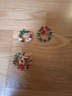 3 juliga broscher med kransmotiv - Säljer tre snygga broscher med julkransmotiv i metall. En har en ren med röda och gröna stenar, en har vita detaljer och grön rosett, och en har en stor röd rosett med klocka. Perfekt för att maxa julstilen! Färgerna är röd, grön, vit och guld.