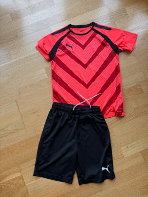 Junior röd och svart Puma t-shirt och svarta Puma shorts  - Sportig t-shirt från Puma i rött med svarta detaljer och grafiskt mönster på framsidan, stl 164. Kortärmad med rund hals och logga på bröstet. Svarta Puma shorts i stl XS.