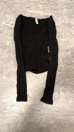 Svart omlottopp med långa ärmar - Svart ribbad omlottopp från hm med lång ärm och v-ringad skärning. Toppens material är mjuk och stretchig bomullsmix, perfekt för en stilren look. Snygg croppad passform som funkar till både jeans och kjol.