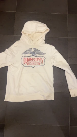 Vit hoodie från Denim & Supply - Vit hoodie från Denim & Supply med stor logga och örnmotiv på bröstet i blått och rött. Tröjan har huva, magficka och långa ärmar. Perfekt för dig som gillar streetwear och vill ha en chill look.