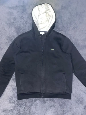 Svart hoodie från Lacoste Sport - Svart hoodie från Lacoste Sport med dragkedja och huva med ljusgrått foder. Klassisk krokodillogga på bröstet och två fickor framtill. Tröjan är långärmad och har en normal passform, perfekt för chill dagar.