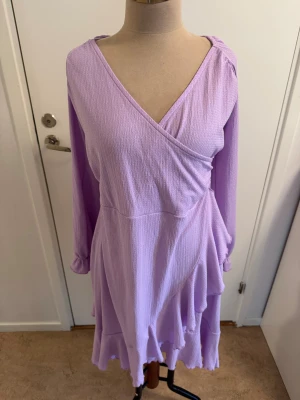 Ljuslila omlottklänning från Bubbleroom - Supersöt ljuslila kortklänning från Bubbleroom med omlott-design, v-ringning och volangdetaljer nertill. Klänningen har långa ärmar med knyt vid ärmslut och är tillverkad i ett lätt, strukturerat tyg som ger en skön känsla. Perfekt för dig som vill sticka ut med en feminin look.