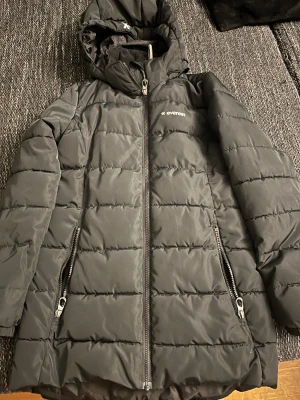Svart pufferjacka från Everest - Svart pufferjacka från Everest med huva och två sidofickor med dragkedja. Jackan har quiltad design och hel dragkedja framtill. Perfekt för kalla dagar och har en stilren look som funkar till det mesta.