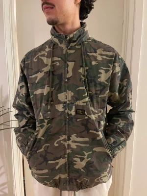 Camouflage vindjacka från Vailent - Säljer en grön och beige camouflage vindjacka från Vailent i storlek L. Jackan har hög krage, snörning vid halsen och dragkedja framtill. Materialet är bomull och nylon, vilket gör den lätt och skön. 