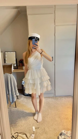Studentklänning  - Supersöt vit kortklänning med omlott-design och lager på lager av volanger. Klänningen har oneshoulder och är tillverkad i ett lätt, skirt material som ger en fluffig och festlig känsla. Perfekt för student eller sommarens speciella tillfällen!