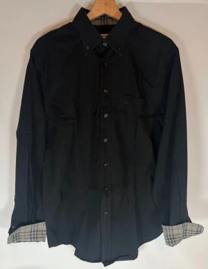 Burberry skjorta - Klassisk svart skjorta från Burberry med rutigt tyg på insidan av manschetterna. Skjortan har knappar hela vägen, bröstficka och lång ärm. Perfekt för dig som gillar stilrena plagg med en twist. Riktigt fint skick och knappast använd. 