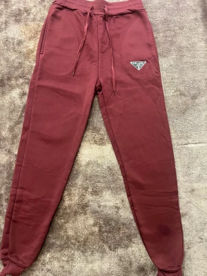 Vinröda mjukisbyxor från Prada - Snygga vinröda mjukisbyxor från Prada med klassisk logga på benet. Byxorna har snörning i midjan, ribbade  vid bensluten och är tillverkade i mjukt bomullsmaterial. Perfekta för chill eller streetwear och har en relaxed passform.