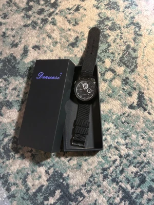 Svart klocka - Snygg svart klocka från Denuasi med rund urtavla och detaljer i vitt. Klockan har ett svart nylonband med metallspänne och flera små visare på urtavlan. Levereras i original ask. Perfekt för dig som gillar stilrena accessoarer.