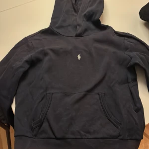Mörkblå hoodie från Polo Ralph Lauren - Snygg mörkblå hoodie från Polo Ralph Lauren med klassisk vit logga broderad på bröstet. Tröjan har huva, känguruficka och lång ärm. Perfekt för chill dagar och enkel att matcha med jeans eller joggers.