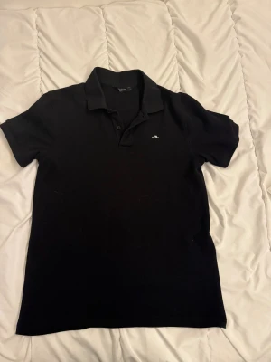 Svart pikétröja från J.Lindeberg - Klassisk svart pikétröja från J.Lindeberg med korta ärmar och liten vit logga på bröstet. Tröjan har krage och knappar framtill, perfekt för en clean och stilren look. Tillverkad i mjuk bomull som känns skön mot huden.