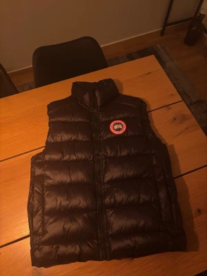 Svart dunväst från Canada Goose - Säljer en svart dunväst från Canada Goose med klassisk logga på bröstet. Västen har hög krage, breda quiltade paneler och dragkedja framtill. Perfekt för lager-på-lager och har en clean, sportig look som funkar till streetwear.