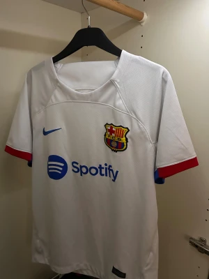 Vit FC Barcelona Nike t-shirt - Säljer en vit FC Barcelona t-shirt från Nike med klubbmärket på bröstet och blå Nike-logga. Spotify-logga i blått framtill. Röd och blå detalj på ärmslut. Perfekt för dig som älskar fotboll och Barca.