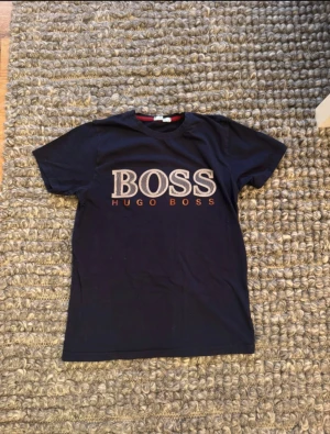 Mörkblå Hugo Boss t-shirt med tryck - Snygg mörkblå t-shirt från Hugo Boss i storlek S. T-shirten har ett stort BOSS-tryck i vitt och orange på bröstet och klassisk rund hals. Materialet är mjuk bomull och modellen är kortärmad, perfekt för en clean och stilren look.