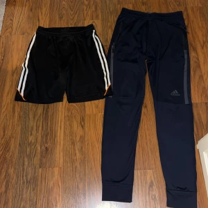 Adidas träningsbyxor & Active Sport shorts - Två par byxor säljes! Adidas mörkblå träningsbyxor med grå detaljer, dragkedjor vid fickorna och smal passform. Active Sport svarta shorts med vita ränder och orange detalj, elastisk midja och snörning. Perfekta för sport och träning.