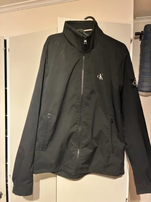 Svart vindjacka från Calvin Klein Jeans - Svart vindjacka från Calvin Klein Jeans med hög krage och dragkedja framtill. Jackan har två fickor med dragkedja och snygga CK-loggor på bröstet och ärmen. Perfekt för dig som gillar stilren och enkel design.