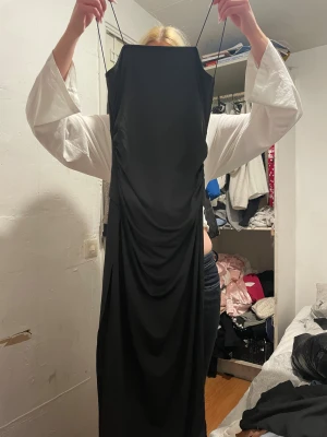 Svart långklänning med smala axelband - Svart långklänning med smala axelband och snygga draperingar både fram och bak. Klänningen har en elegant, figurnära siluett och är tillverkad i ett mjukt, stretchigt material som ger en skön passform. Perfekt för dig som gillar minimalistisk och stilren design.