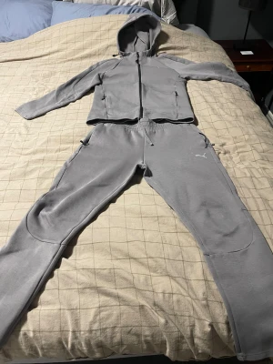 Grå mjukisbyxor från Puma - Säljer ett par grå mjukisbyxor från Puma med klassisk logga på benet. Byxorna har normal passform, elastisk midja med snörning och praktiska fickor med dragkedja. Perfekta för chill eller träning, tillverkade i mjukt syntetmaterial.