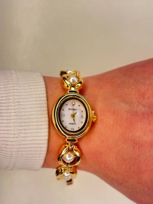 Guldfärgad pärlklocka från Homdy - Snygg guldfärgad armbandsklocka från Homdy med oval urtavla i vitt och små glittrande stenar som timmarkeringar. Länken har dekorativa pärlor infattade i guldfärgade länkar, vilket ger en lyxig och elegant vibe. Quartz-urverk och smidig låsning. Det medföljer en klick justerare. 