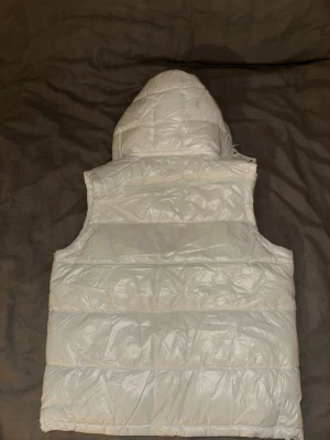 Vit Moncler dunväst med huva - Säljer en snygg vit dunväst från Moncler med huva och dragkedja framtill. Västen har quiltad design, två fickor med dragkedja och klassisk Moncler-logga på bröstet. Insidan har en cool serietidningsetikett. Perfekt för lager-på-lager under kyliga dagar.