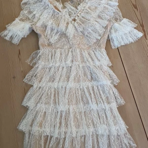 Vit spetsklänning från Malina - Superfin vit spetsklänning från Malina med volanger i flera lager och v-ringad omlott. Klänningen har korta ärmar med volangdetaljer och är tillverkad i ett tunt, elegant spetsmaterial. Använd 1 gång på studenten💕 har en defekt, men inget som påverkar