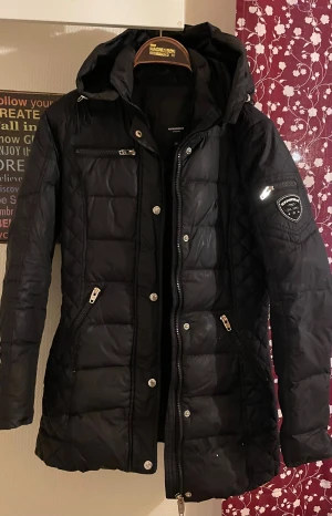 Svart quiltad pufferjacka Rockandblue - Snygg svart pufferjacka från Rockandblue med quiltade detaljer och huva. Jackan har flera dragkedjeförsedda fickor, silverfärgade knappar och Rockandblue-logga på ärmen. Perfekt för kalla dagar med sin långa passform och stilrena look.Är använd så det finns några ”skavanker ” Men inget som stör!