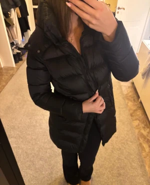 Svart lång pufferjacka med huva - Säljer en svart lång pufferjacka med huva och dragkedja från BomBoogie. Jackan är vadderad och har en clean look med diskreta sömmar och en figurnära passform. Nypris: 4800kr, pris kan diskuteras!!!