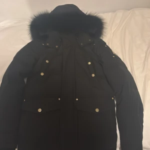 Svart Big Ridge Parka från Moose Knuckles - Säljer en svart Big Ridge Parka från Moose Knuckles med fluffig päls runt huvan, guldfärgade knappar och ikonisk logga på ärmen. Jackan har flera fickor, lång ärm och är perfekt för kalla dagar. Stilsäker och rejäl design med snygga detaljer. Den är storlek XS men passar S. Pris är inte hugget i sten