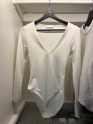 Vit långärmad bodysuit med v-ringning - Säljer en stilren vit bodysuit från H&M med djup v-ringning och lång ärm. Modellen är tight och sitter snyggt mot kroppen. Materialet är mjuk bomull med lite stretch. Aldrig använt. 
