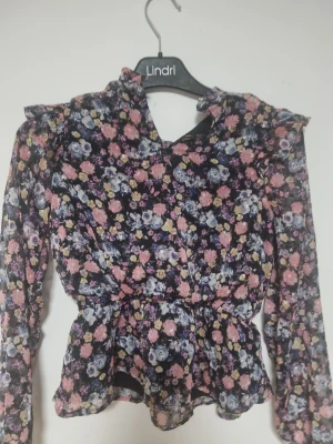 Blommig blus från Lindex Kids - Supersöt blommig blus från Lindex Kids i storlek 140. Blusen har volangdetaljer vid axlarna, lång ärm och knäppning bak i nacken. Färgerna går i svart, rosa, lila, gul och blå. Perfekt för dig som gillar romantiska och färgglada plagg!