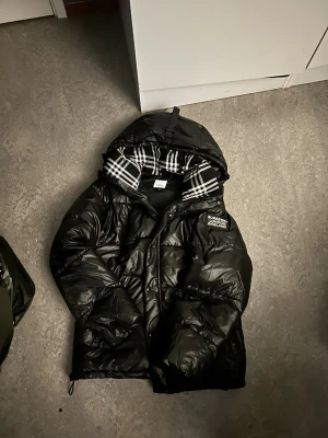 Svart pufferjacka från Burberry L - Svart pufferjacka från Burberry med stor huva och klassiskt rutigt foder i svart och vitt. Jackan har en tydlig Burberry London England-patch på bröstet och dragkedja framtill. Yttermaterialet är bomull och fodret är också bomull. Perfekt för kalla dagar.