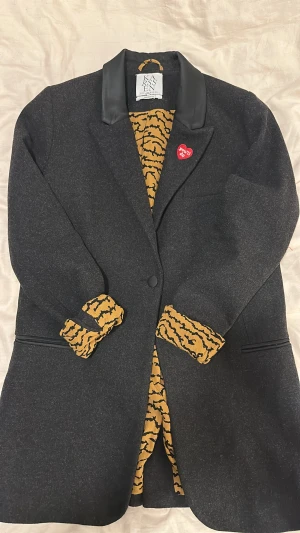 Svart kavaj från Zoe Karssen M - Svart kavaj med coola detaljer från Zoe Karssen. Kragen är i svart skinnimitation och insidan har ett tigerinspirerat mönster i gult och svart. På bröstet sitter ett rött hjärta med texten 'you'll do'. Kavajen har knappar och fickor med svarta detaljer.