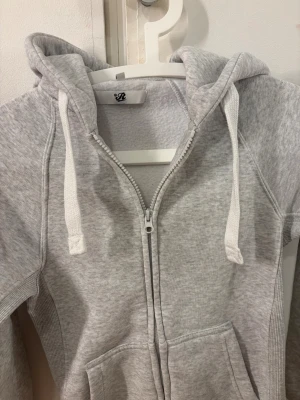 Ljusgrå hoodie från Russemerch XXS - Ljusgrå hoodie från Russedress i storlek XXS. Tröjan har dragkedja framtill, huva med snörning och två fickor. Mjuk bomullsmix och ribbade muddar vid ärmslut och nederkant. Diskret Russedress-logga på ärmen. Perfekt för chill dagar.
