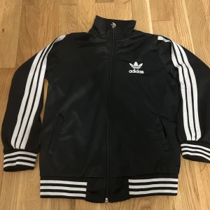Svart Adidas vindjacka med vita ränder - Klassisk svart Adidas vindjacka med vita ränder längs ärmarna och broderad logga fram och bak. Jackan har dragkedja, ribbade muddar och krage. Perfekt för en sportig look och enkel att matcha till det mesta.