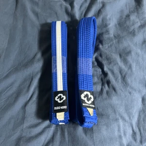 Blått kampsportsbälte Budo-Nord - Två blå kampsportsbälten från Budo-Nord, ett helblått och ett med vit rand längs mitten. Bältena är tillverkade i kraftig bomull och har klassisk brottningsstruktur. Perfekt för judo, karate eller annan budo-träning.
