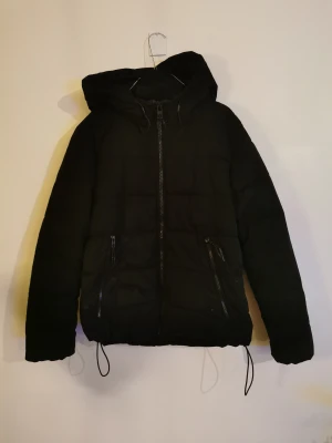 Svart pufferjacka med huva - Kort svart "dunjacka"/ täckjacka. Huva som sitter fast. Dragsko i midja för att kunna justera passformen. Jackan har två sidofickor med dragkedjor.  Något sliten i tyget vid arm och vid ficka annars bra skick (se bild). Det ser värre ut på bild än verklighet.  Varm och skön sål perfekt för kalla dagar. Stl M från FB Sister (New yorker) 