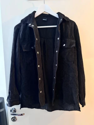Svart manchester overshirt från Gina Tricot - Svart overshirt i mjuk manchester från Gina Tricot. Jackan har klassisk krage, långa ärmar och silverfärgade knappar framtill samt på fickorna. Perfekt att slänga över en hoodie eller t-shirt för en chill look.