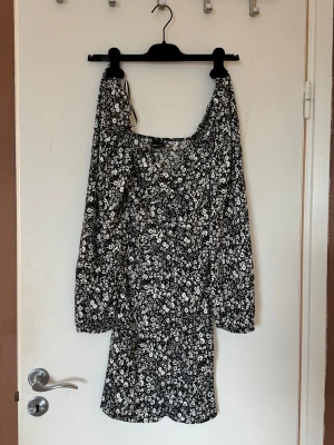 Svartvit blommig kortklänning - Supersöt svartvit kortklänning med blommigt mönster och långa puffärmar. Klänningen har en lätt rynkad byst och markerad midja som ger en smickrande siluett. Perfekt för dig som gillar en feminin och trendig stil.