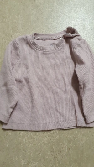 Tröja - Säljer en ljuslila långärmad tröja från I Love Eco i ribbad bomull. Tröjan har praktiska tryckknappar vid axeln för enkel påklädning och en rund halsringning. Perfekt för dig som gillar mjuka och sköna basplagg med enkel design.