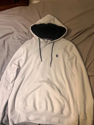 Vit hoodie från US Polo Assn M - Vit hoodie från US Polo Assn med svart insida på huvan och svarta snören. Klassisk broderad logga på bröstet och stor magficka framtill. Skön och avslappnad passform, perfekt för chill dagar. Materialet är mjuk bomull.