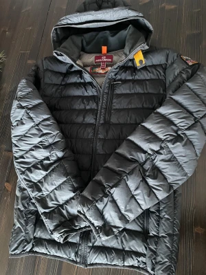 Parajumpers -Svart lättvikts dunjacka  - Svart lättvikts dunjacka (Herrmodell)  från Parajumpers med huva. Jackan är vadderad med 90% dun och 10% fjäder. Yttermaterialet är både vattenavvisande och vindtätt. Framtill finns två fickor med dragkedja. Små ljusare, knappt synbara, streck under märket (går ev. bort i tvätten), kan ses på bild 3. Köpt på Kids Brand Store, nypris 4800kr. Storlek YL (Youth Large), passar vuxen Small. 