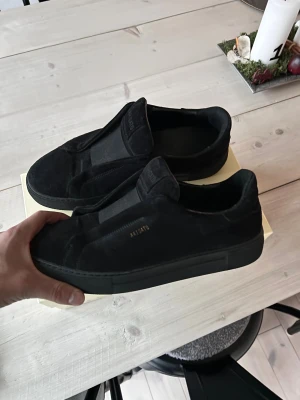 Svarta sneakers Axel Arigato mocka - Svarta sneakers från Axel Arigato i mjuk mocka med diskret logga i guld på sidan. Snygg minimalistisk design utan snörning, med elastisk panel över foten för enkel påtagning. Platt sula och rund tå ger en clean och modern look. Obs modellen är discontinued 