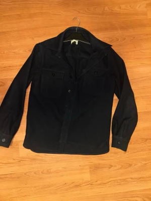 Svart overshirt - Pike Bros - Säljer en svart overshirt i ull med klassisk krage och två stora bröstfickor med knappar. Jackan har långa ärmar med knapp vid mudden och stängs med knappar framtill. Perfekt lager-på-lager-plagg för kyligare dagar. 9/10 behövs endast rolla lite pga småhår o ludd. LÄGG GÄRNA BUD