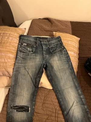 G-Star Raw blå jeans med patch - Säljer ett par blå G-Star Raw jeans med slitningar och patchad detalj på ena benet. Jeansen har raka ben, stora bakfickor med dragkedja och coola sömmar