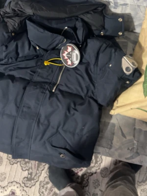 Mörkblå vinterjacka från Moose Knuckles - Säljer en mörkblå vinterjacka från Moose Knuckles med ikonisk metallogo på ärmen och snygga detaljer. Jackan har hög krage, dragkedja och flera fickor framtill. Perfekt för kalla dagar och riktigt stilren look.