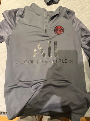 Grå PSG Nike Dri-Fit träningströja - Snygg grå träningströja från Nike med Paris Saint-Germain-logga på bröstet och röd Nike Swoosh på ärmen. Tröjan har halv dragkedja och är tillverkad i Dri-Fit material som andas. Perfekt för fotbollsträning eller chill. Stort 'ALL' tryck på magen.