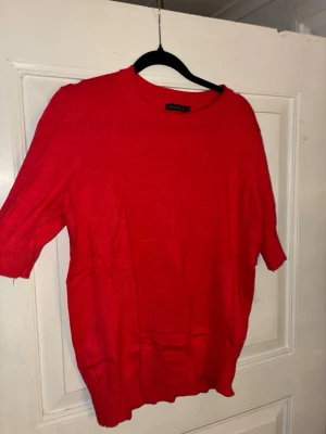 Röd stickad tröja från Stockh LM - Röd stickad tröja från Stockh LM med rund halsringning och korta ärmar. Tröjan har en enkel design med ribbade muddar och en liten metallring i nacken som detalj. Perfekt att styla med jeans eller kjol.